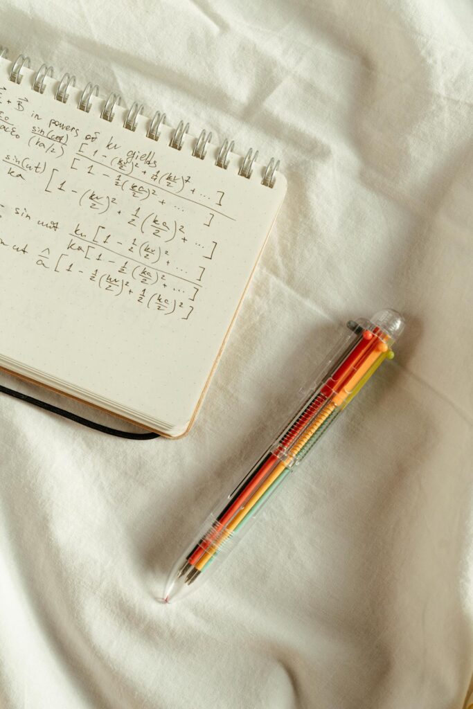 Image of a multi-colored pen set against a white sheet with a notebook showing equations on it. Photo by cottonbro studio: https://www.pexels.com/photo/notepad-and-a-pen-5184945/